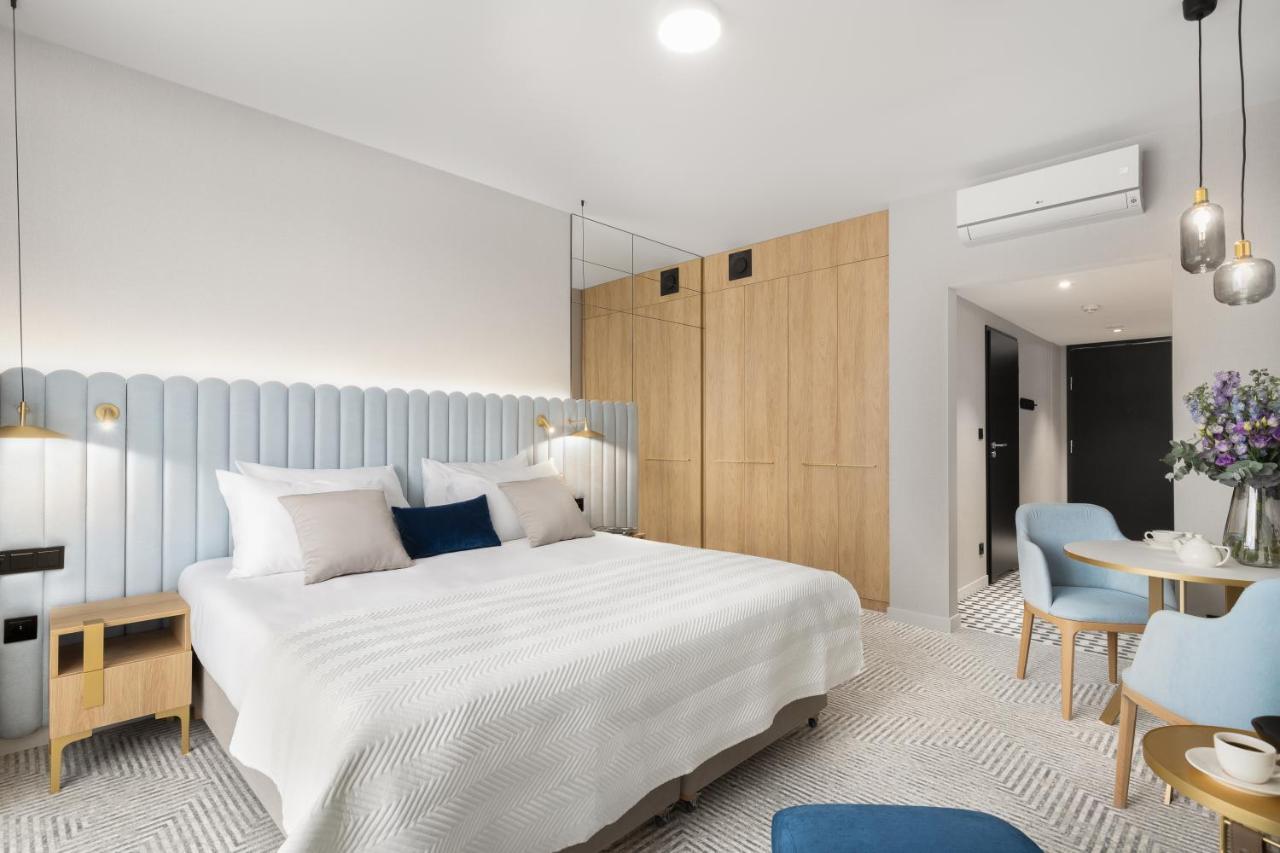 Royal Tulip Warsaw Centre Aparthotel Exterior foto The photo shows a modern hotel room featuring a spacious layout. There's a large double bed with a light-colored bedspread, accented by decorative pillows. The headboard is light blue, providing a soft contrast to the wooden walls. On one side of the
