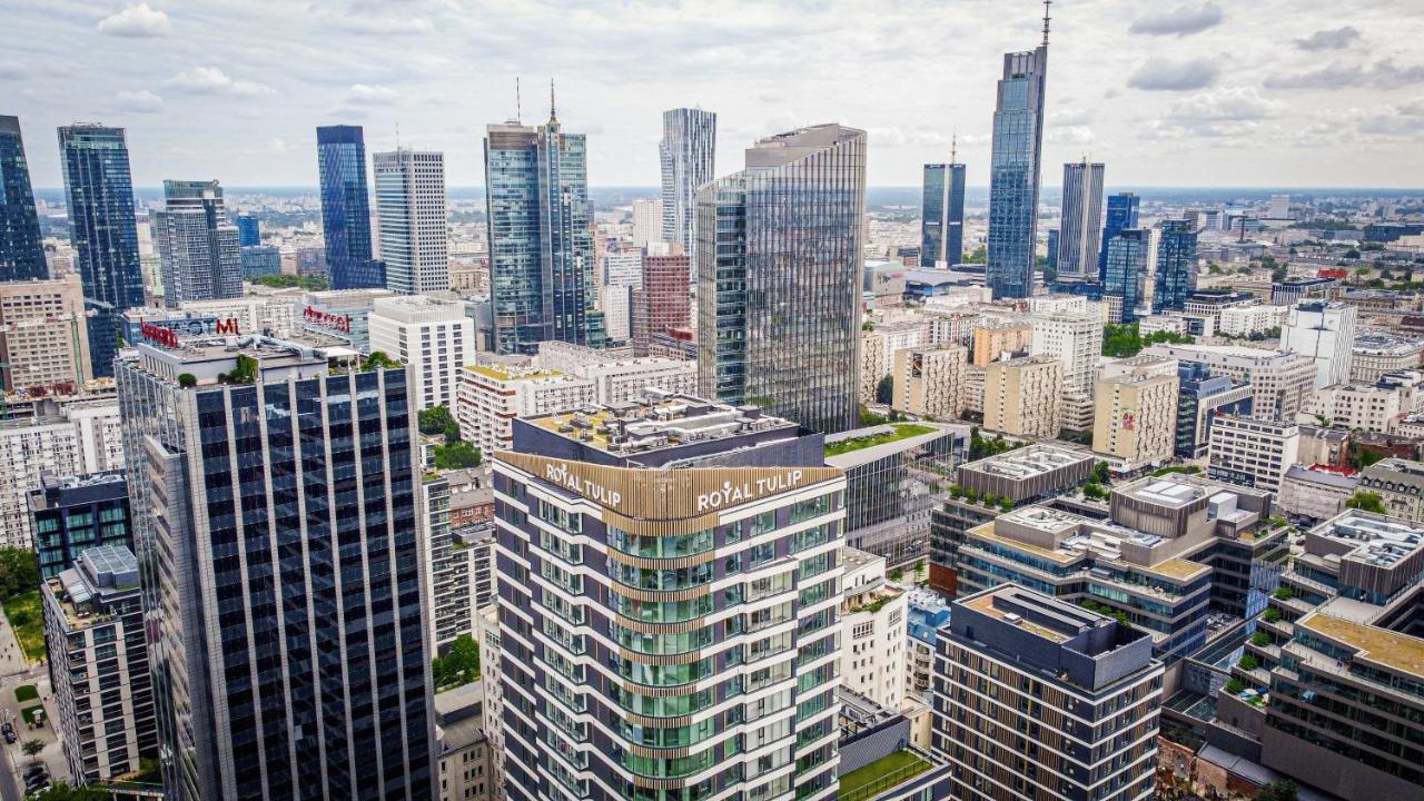 Royal Tulip Warsaw Centre Aparthotel Exterior foto The photo shows a panoramic view of a modern city skyline. In the foreground, there are several tall buildings with contemporary architectural designs. One building prominently features the name "Royal Tulip." The background is filled with numerous s