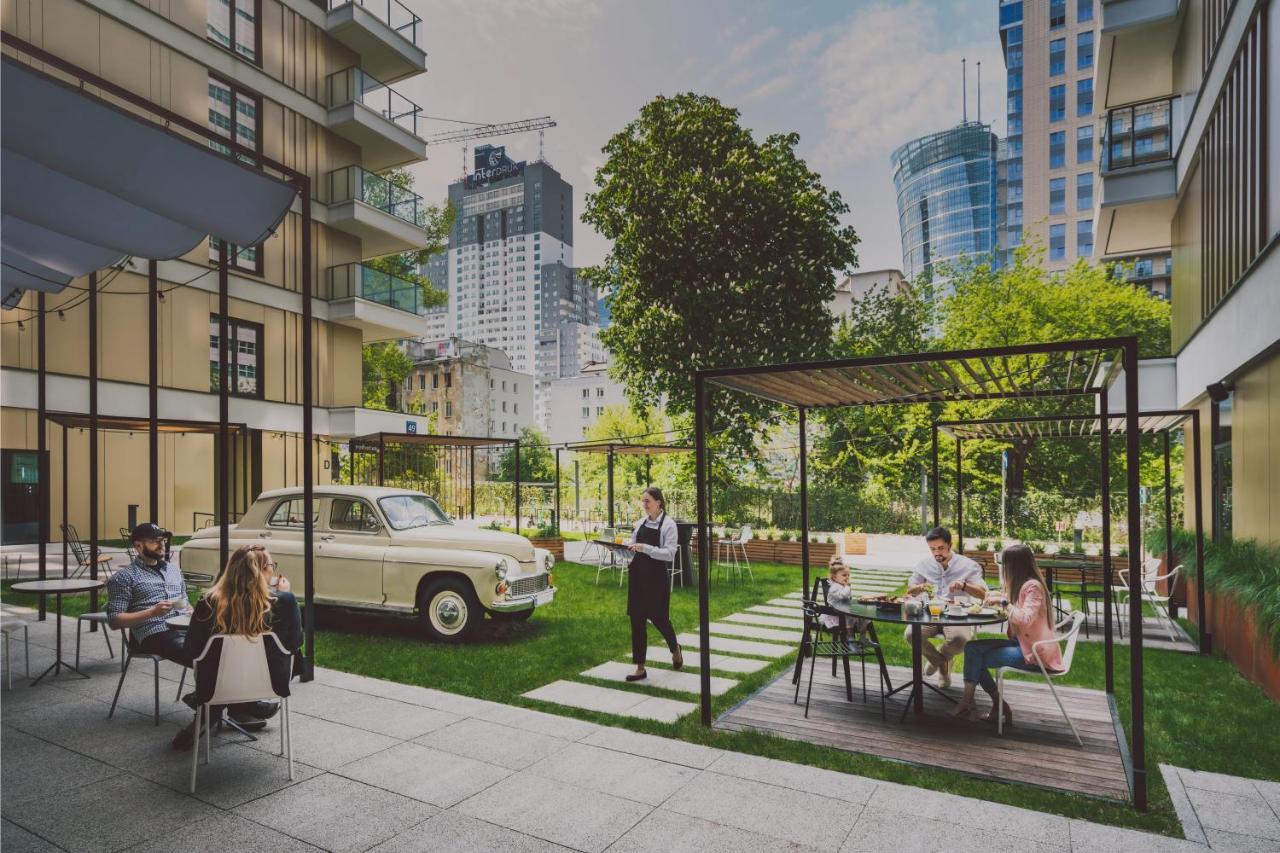 Royal Tulip Warsaw Centre Aparthotel Exterior foto The photo depicts a modern urban setting featuring a green courtyard. In the foreground, there are several people sitting at tables, enjoying a meal or drinks. A couple of individuals are lounging, while others appear to be dining. 

In the backgroun