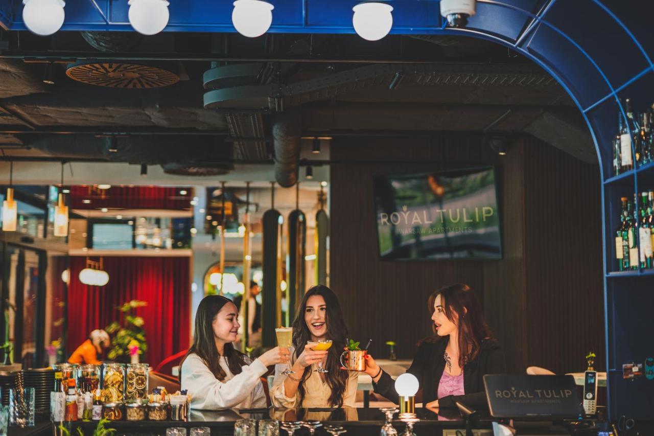 Royal Tulip Warsaw Centre Aparthotel Exterior foto The photo depicts three women gathered around a bar, enjoying drinks. They are smiling and engaged in conversation, exuding a festive and cheerful atmosphere. The bar has a modern design, with colorful decor and lighting. In the background, there is 