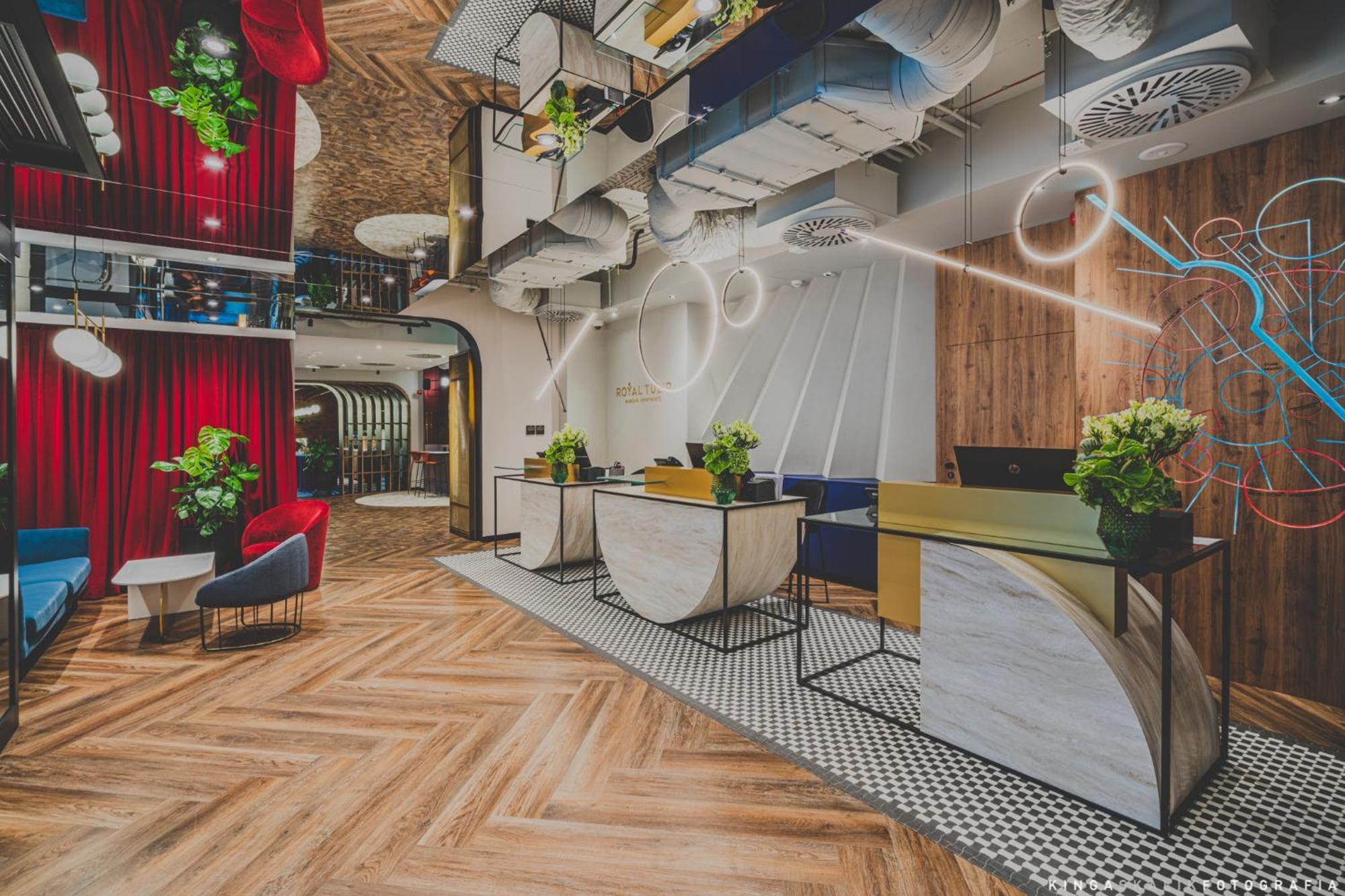 Royal Tulip Warsaw Centre Aparthotel Exterior foto The photo shows a modern hotel lobby with a stylish design. It features a reception area with two main desks, each adorned with plants. The floor has a patterned wooden design, and there are colorful chairs placed around the space. The ceiling is dec