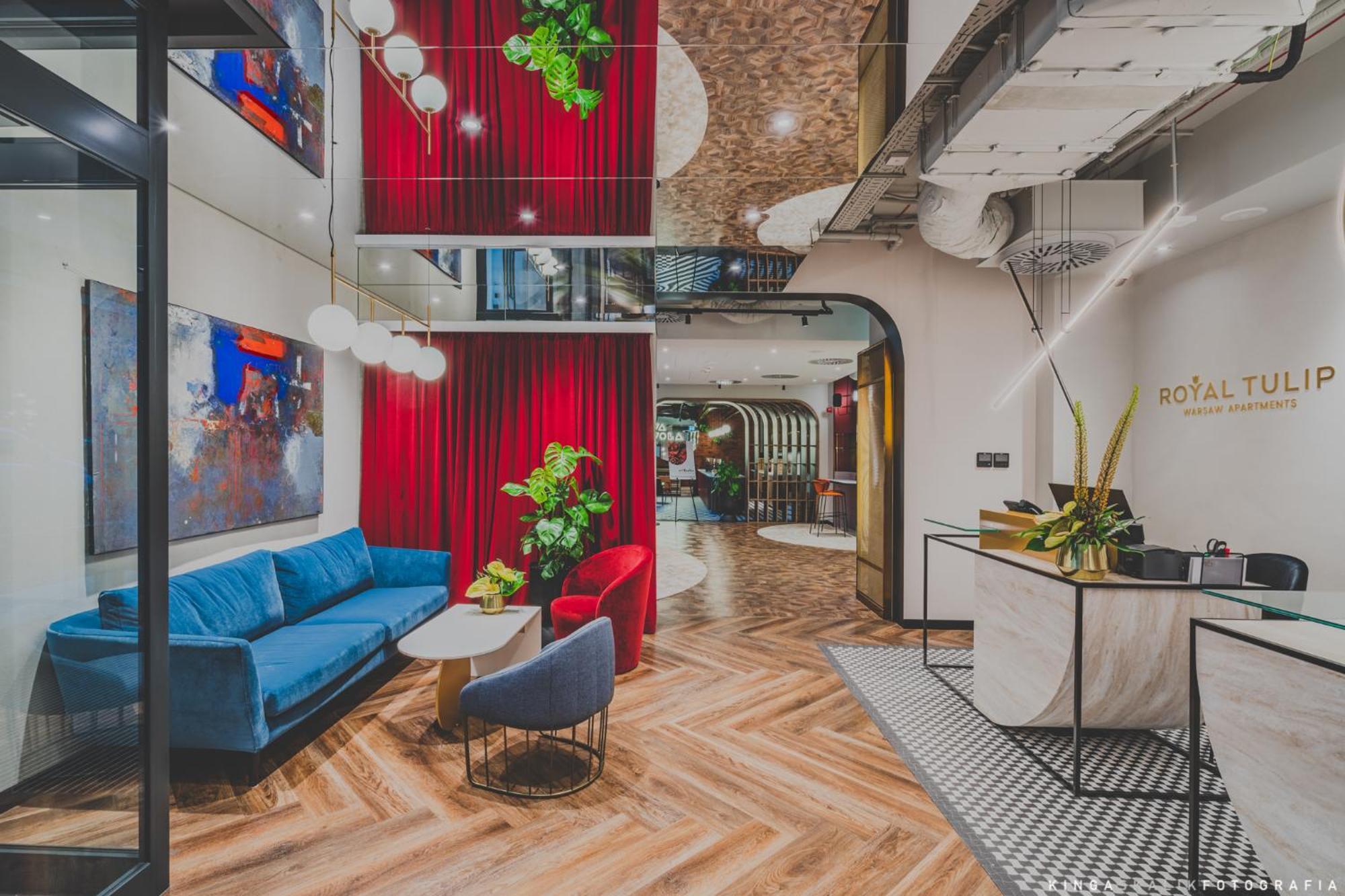 Royal Tulip Warsaw Centre Aparthotel Exterior foto The photo showcases a modern interior space, likely a lobby or lounge area. It features a mix of textures and colors, including a blue couch and red accent chairs. Behind the seating area, there are vibrant red curtains and greenery that adds a touch
