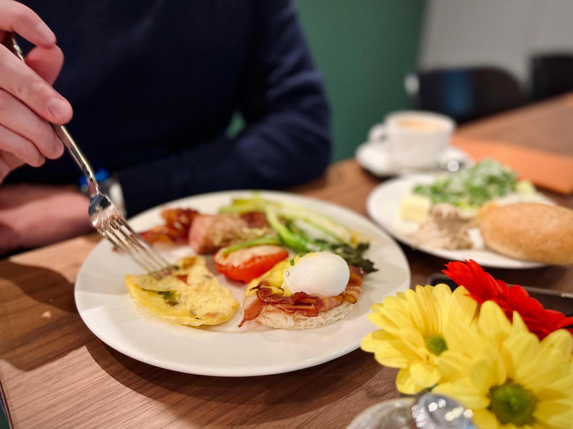 Royal Tulip Warsaw Centre Aparthotel Exterior foto The photo displays a breakfast plate with various items. There's an omelet, a poached egg, bacon, sliced tomatoes, and some greens. A hand is reaching for the food with a fork. Additionally, there are flowers on the table and a small cup of coffee. T