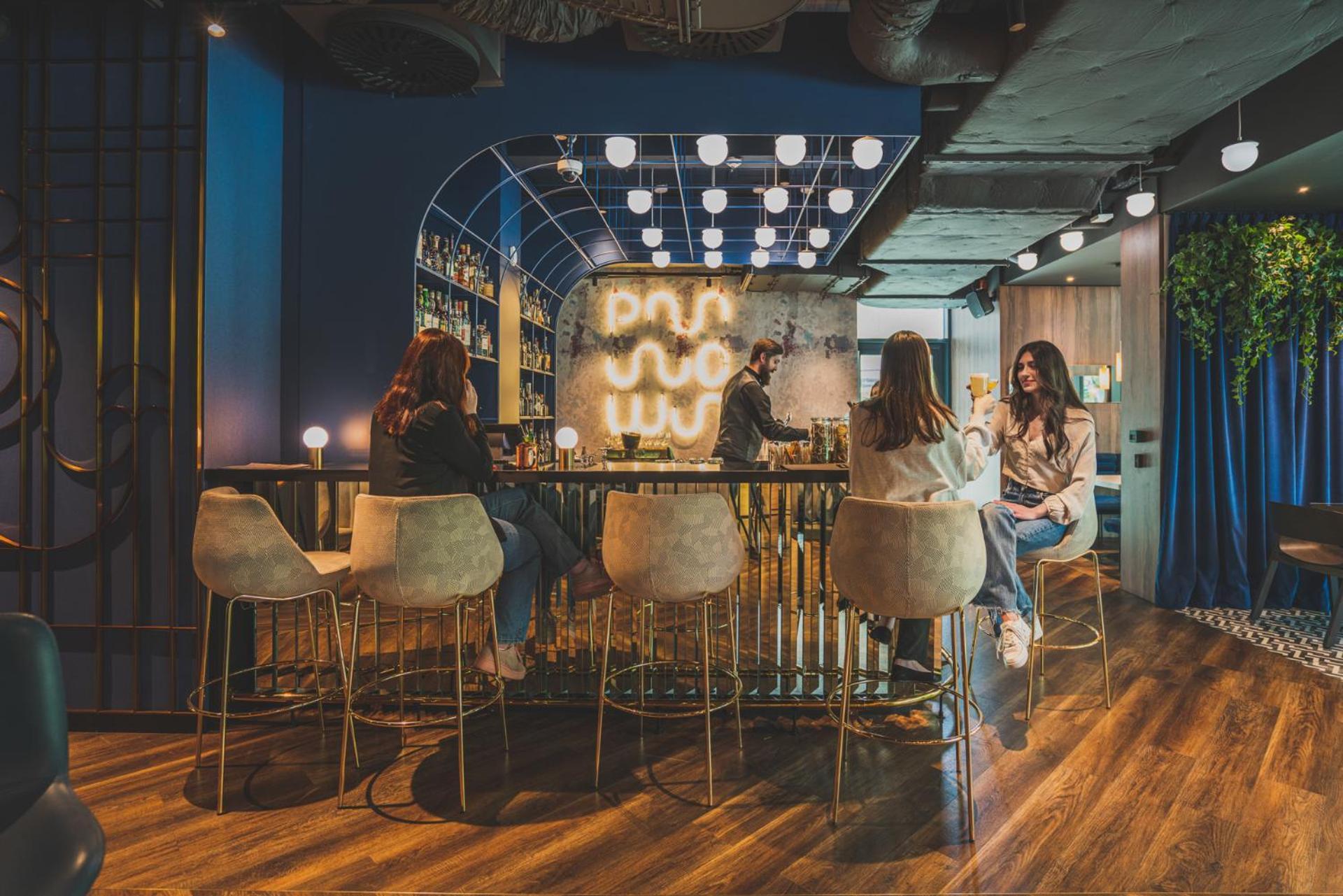Royal Tulip Warsaw Centre Aparthotel Exterior foto The photo depicts a modern, stylish lounge or bar interior. There are several people sitting at a curved bar with high-back stools, engaged in conversation. The bar area is well-lit with decorative lighting that creates a warm atmosphere. In the back