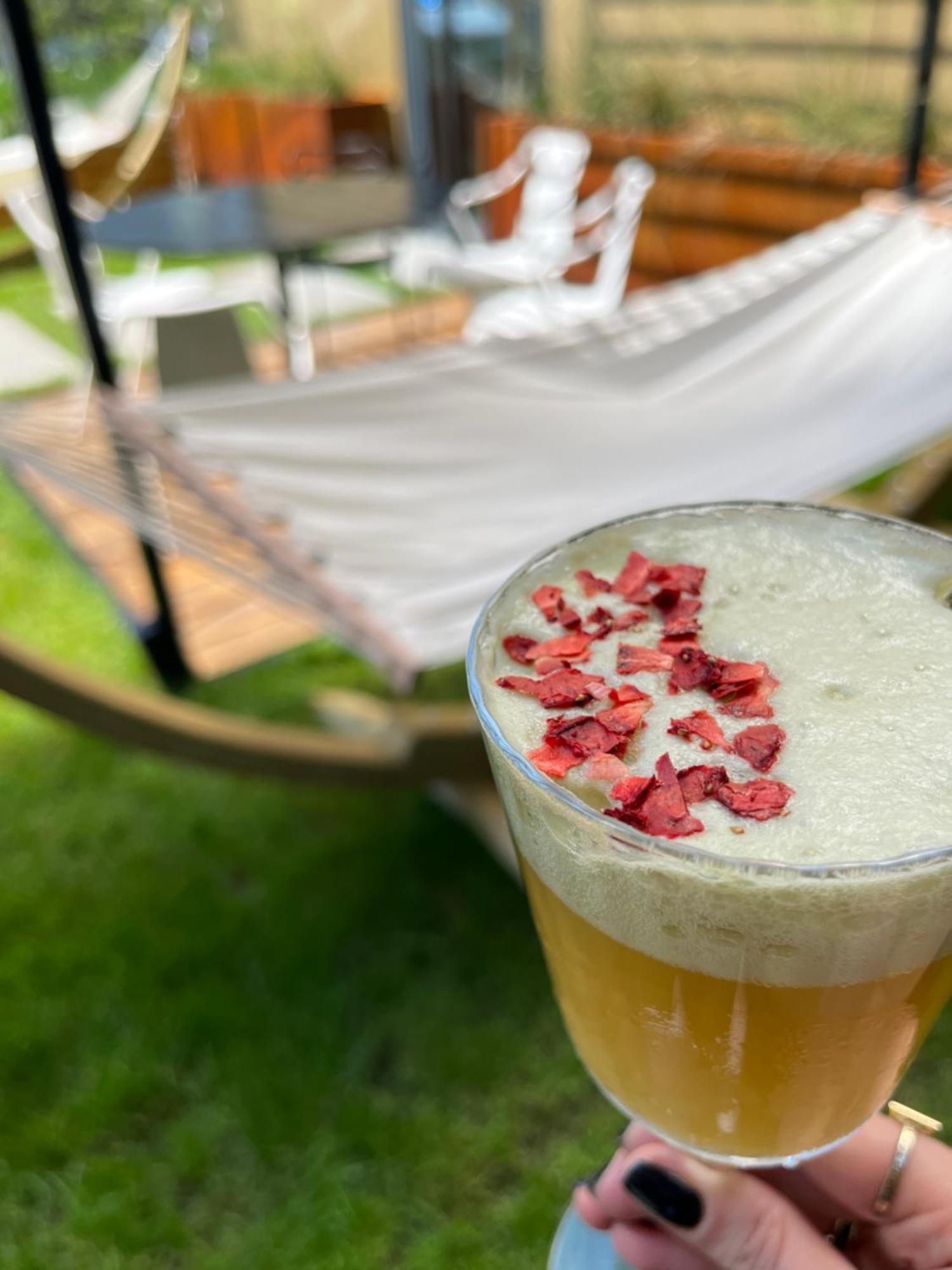 Royal Tulip Warsaw Centre Aparthotel Exterior foto The photo shows a close-up of a drink in a glass, topped with small pieces of red petals or dried flowers. The drink appears to have a frothy white layer above a golden-yellow liquid. In the background, there is a hammock set up on the grass, indicat