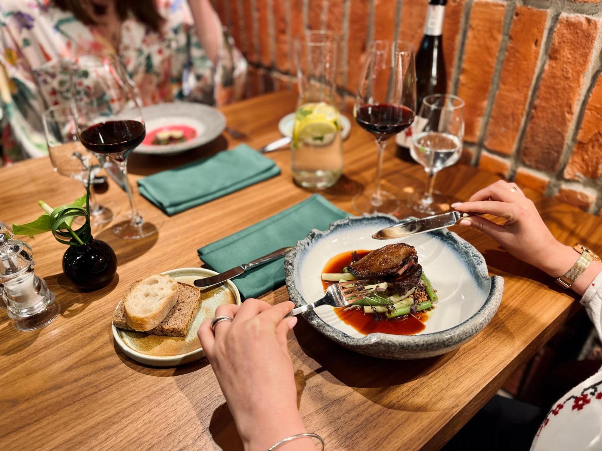 Royal Tulip Warsaw Centre Aparthotel Exterior foto The photo shows a dining scene at a restaurant. There’s a beautifully presented plate of food in the foreground, featuring a piece of meat placed on a bed of greens, garnished with sauce. A person's hand is holding a fork and knife, ready to cut into
