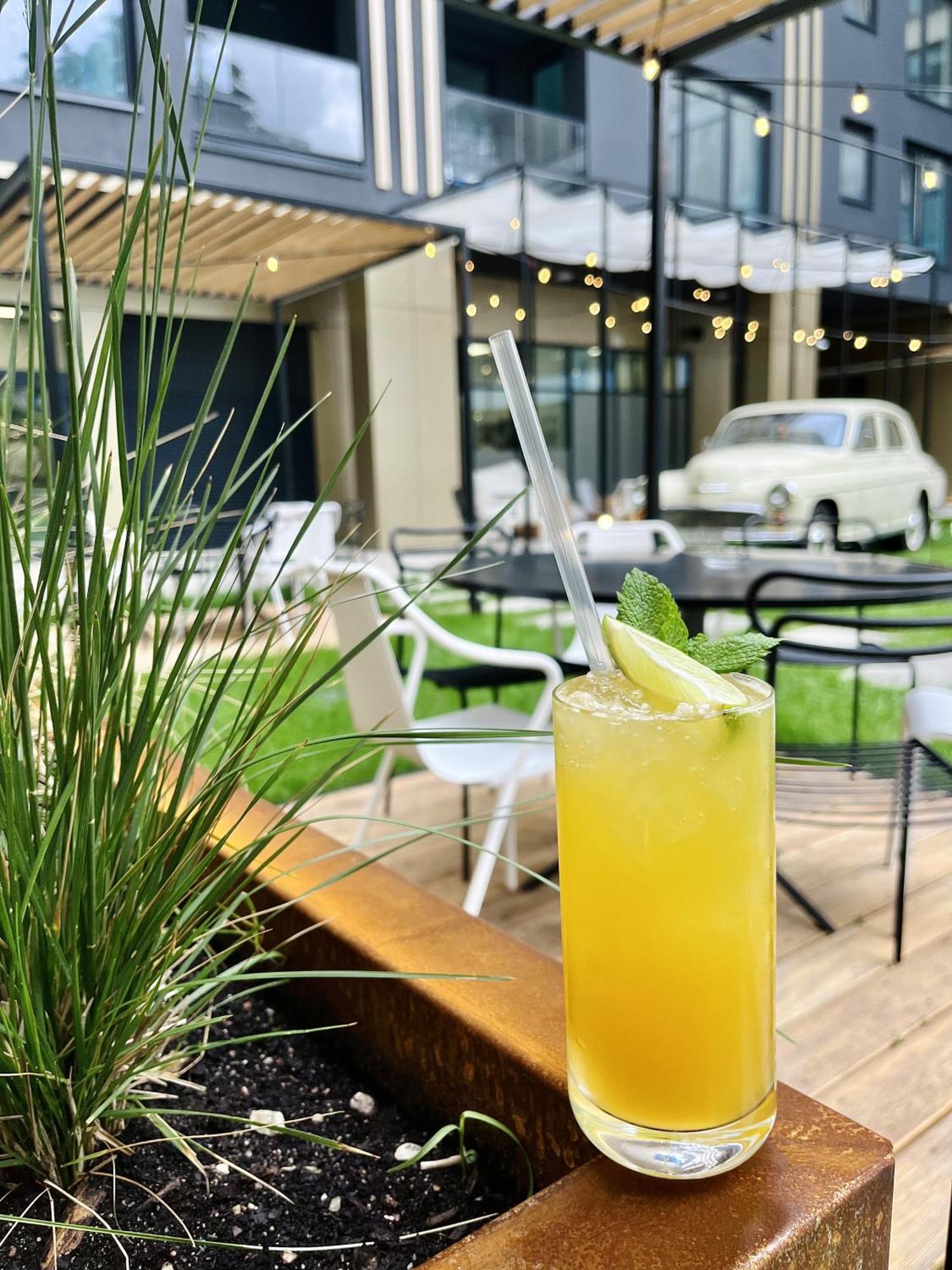 Royal Tulip Warsaw Centre Aparthotel Exterior foto The image shows a refreshing cocktail placed prominently in the foreground, featuring a vibrant yellow color, topped with a slice of lime and a sprig of mint. There is a clear straw inserted into the drink. In the background, there are outdoor seatin