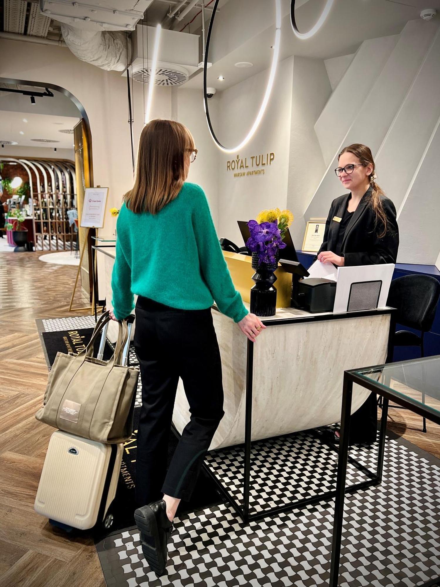 Royal Tulip Warsaw Centre Aparthotel Exterior foto The image shows a hotel reception area. A woman wearing a green sweater and black pants is standing at the reception desk, engaging with a staff member. The staff member, who is smiling and wearing a name tag, is standing behind the desk. There's a b